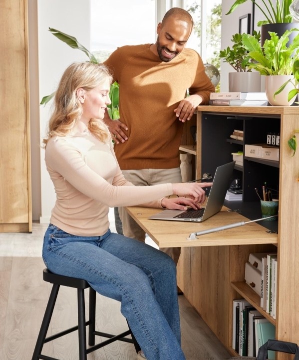 Wohnzimmer mit Mann und Frau vor einer Kommode mit Schreibtischaufsatz und Büchern.