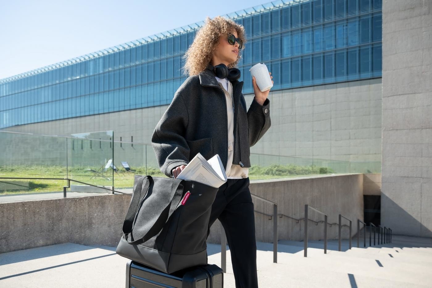 Person mit lockigem Haar mit Rollkoffer und Reisenthel Travelshopper in schwarz vor einem Gebäude mit Glasfront.