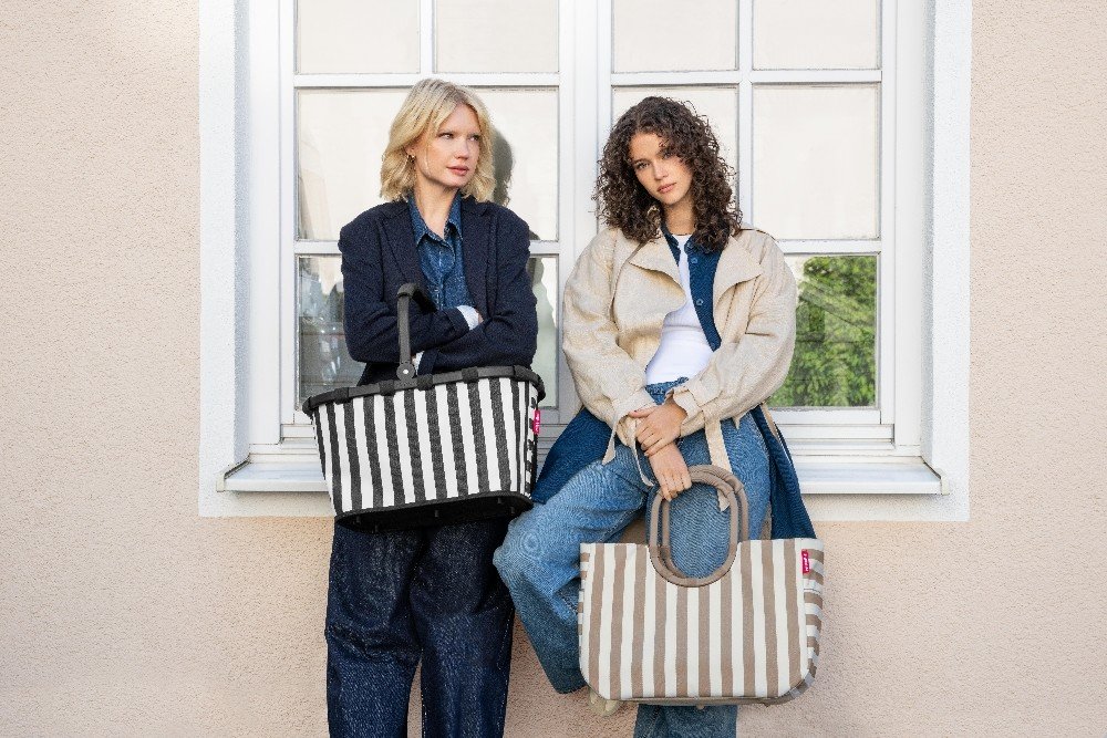 Zwei Frauen jeweils mit sommerlich gestreiftem Reisenthel Carrybag und großem Loopshopper am Arm, die vor einer Häuserwand mit Fenster stehen.