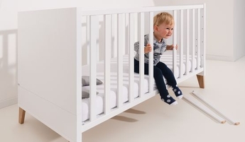 Weißes Gitterbett mit zwei entfernten Schlupfsprossen und Kleinkind, das  auf der Bettkante sitzt